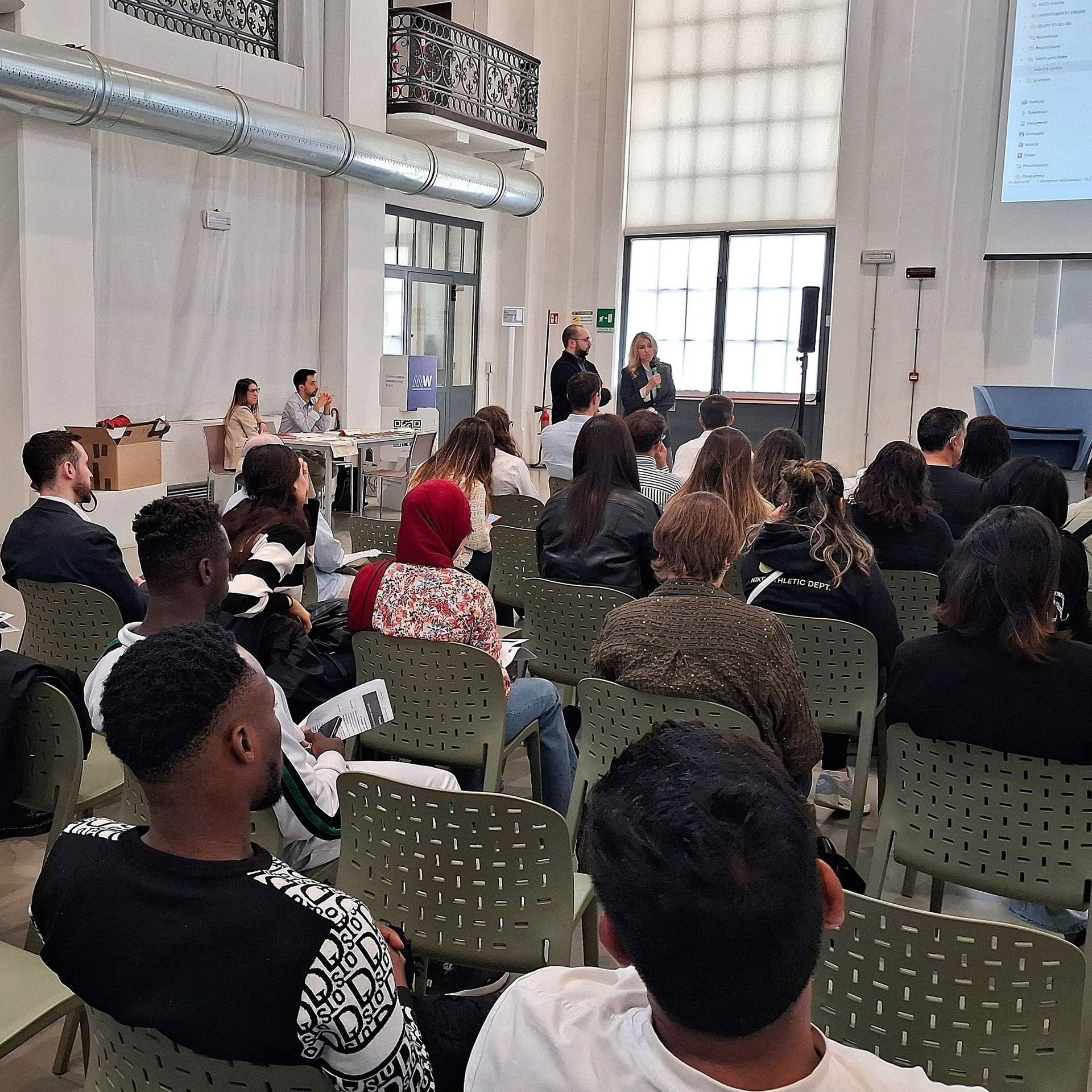 Presentazione di una conferenza. Relatori sul palco, pubblico seduto su sedie, all'interno.