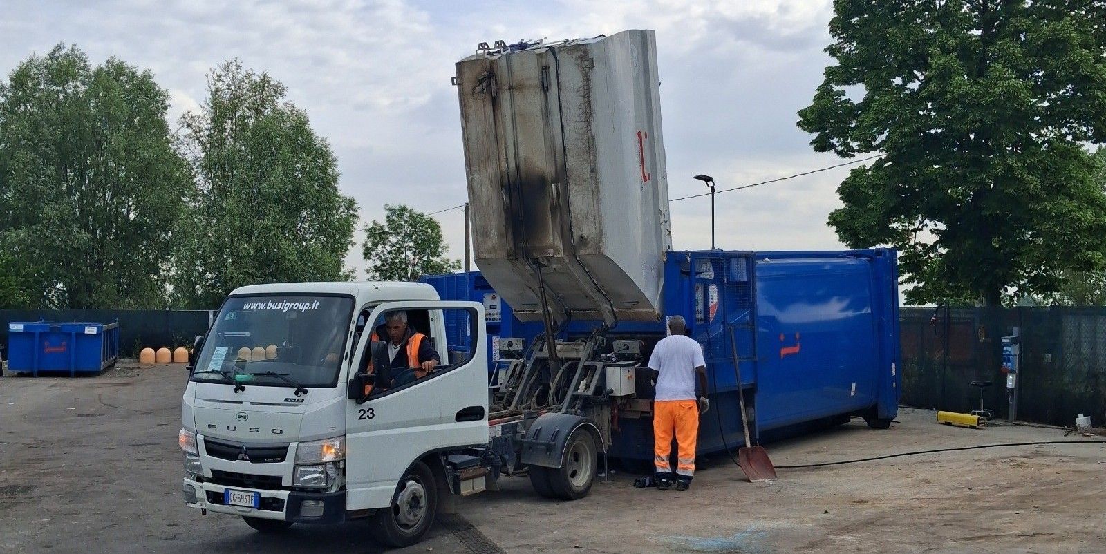 Un camion della spazzatura scarica rifiuti in un grande contenitore blu. Due operai sono presenti all'aperto.
