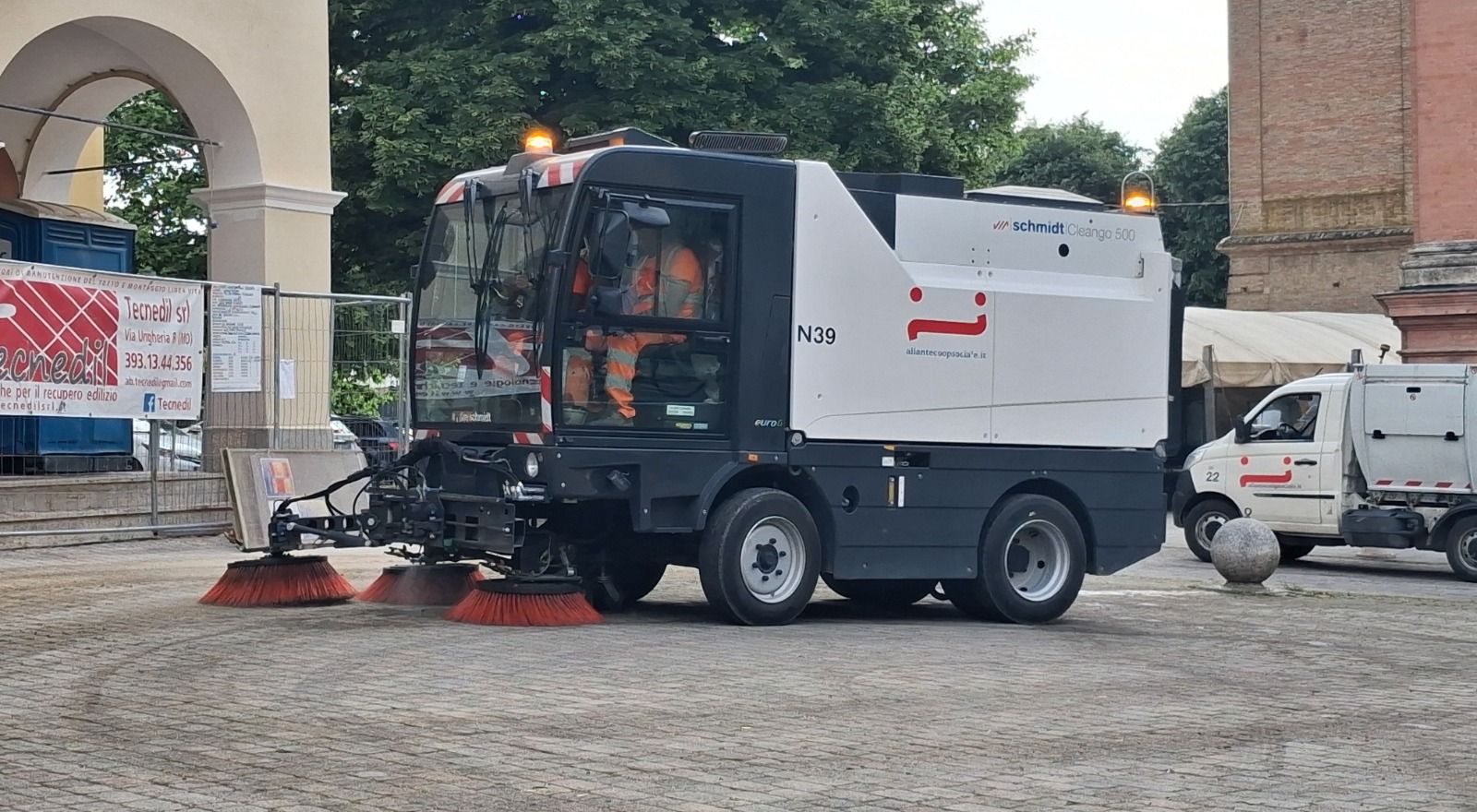 Spazzatrice stradale che pulisce un'area pavimentata di fronte a un edificio. La spazzatrice è grigia e bianca.