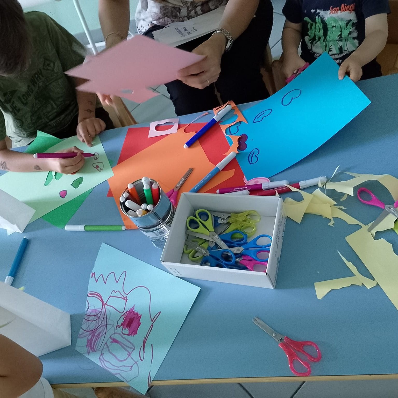 Bambini e un adulto lavorano su un tavolo blu, utilizzando carta, pennarelli e forbici.