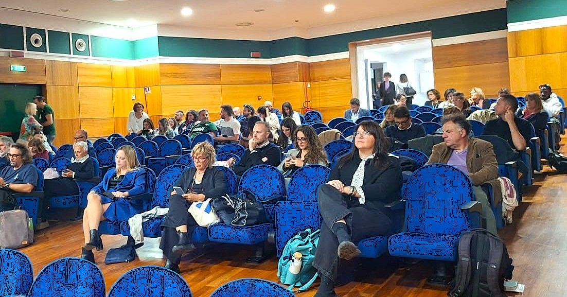Il pubblico è seduto su sedie blu, in una grande sala con pannelli in legno. La gente ascolta, guardando avanti.
