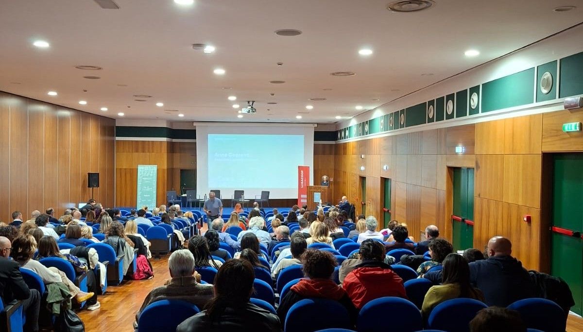 Pubblico in una sala conferenze, di fronte a un palco con altoparlanti e uno schermo.