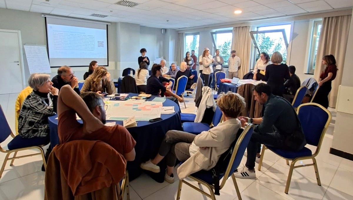 Le persone si sono radunate per una conferenza attorno a dei tavoli, con uno schermo di proiezione visibile sullo sfondo.