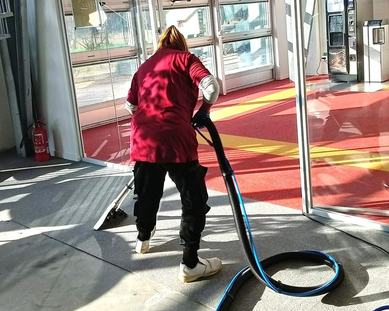 Persona che passa l'aspirapolvere sul pavimento all'interno di un edificio con una grande porta a vetri. Camicia rossa, pantaloni neri, scarpe bianche.