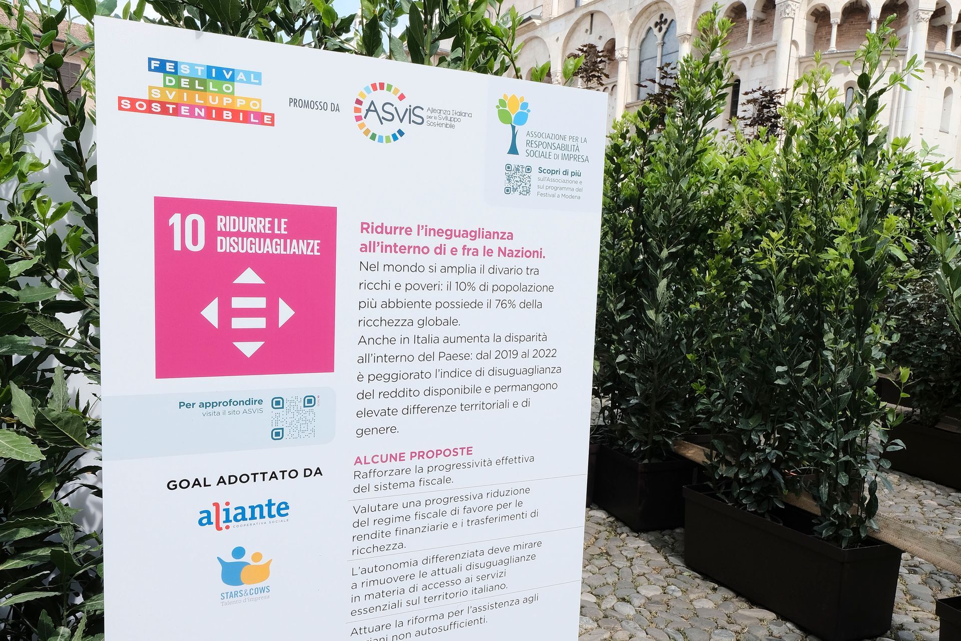 Cartello con l'icona dell'SDG 10, piante ed edificio sullo sfondo. Promuovere la riduzione delle disuguaglianze.