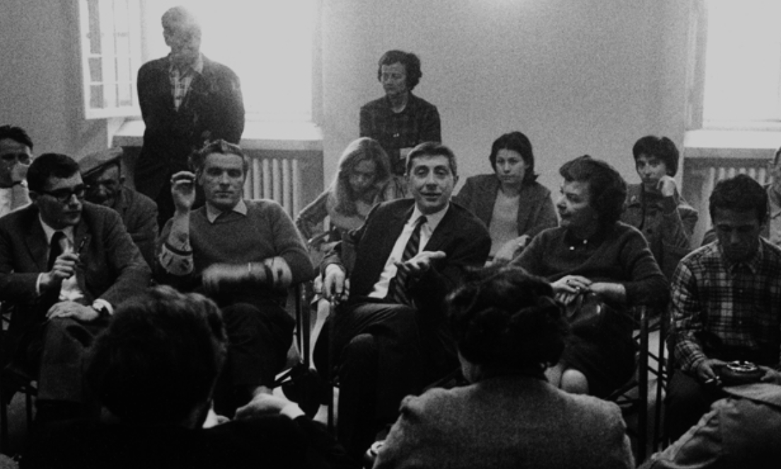 Basaglia e altre persone sedute in una stanza, alcune guardano un uomo che parla, altre ascoltano. Ambientazione interna, bianco e nero.