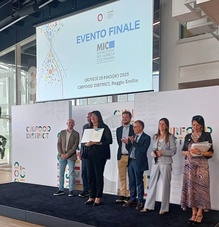 Persone sul palco durante un evento, mentre ricevono un premio. Sullo sfondo un logo e la scritta 