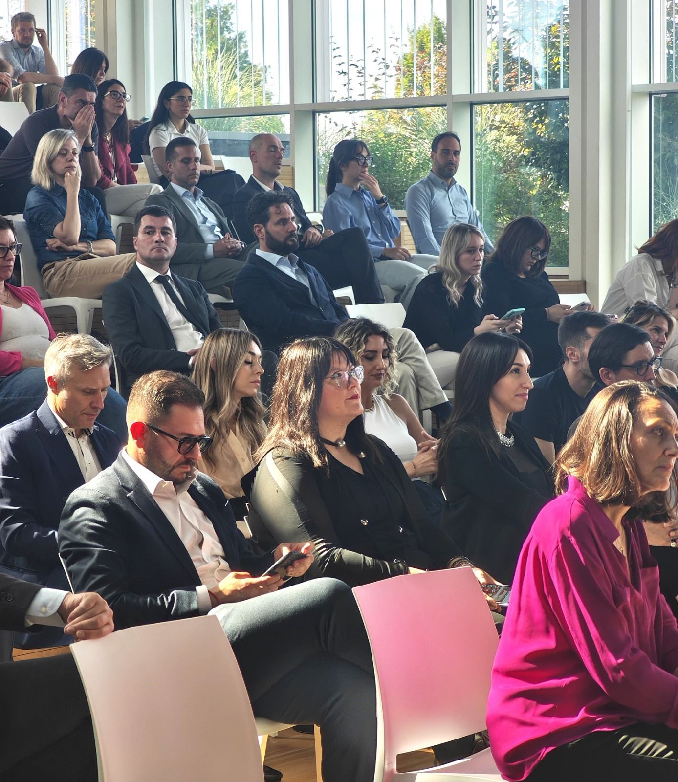 Il pubblico è seduto in una sala illuminata dal sole, molti guardano avanti. Molti indossano abiti eleganti e colori scuri. Sedie rosa e bianche.