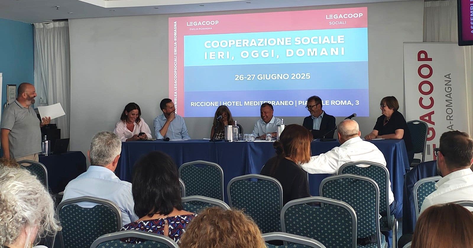 Tavola rotonda: relatori al tavolo, pubblico in primo piano, striscione con la scritta