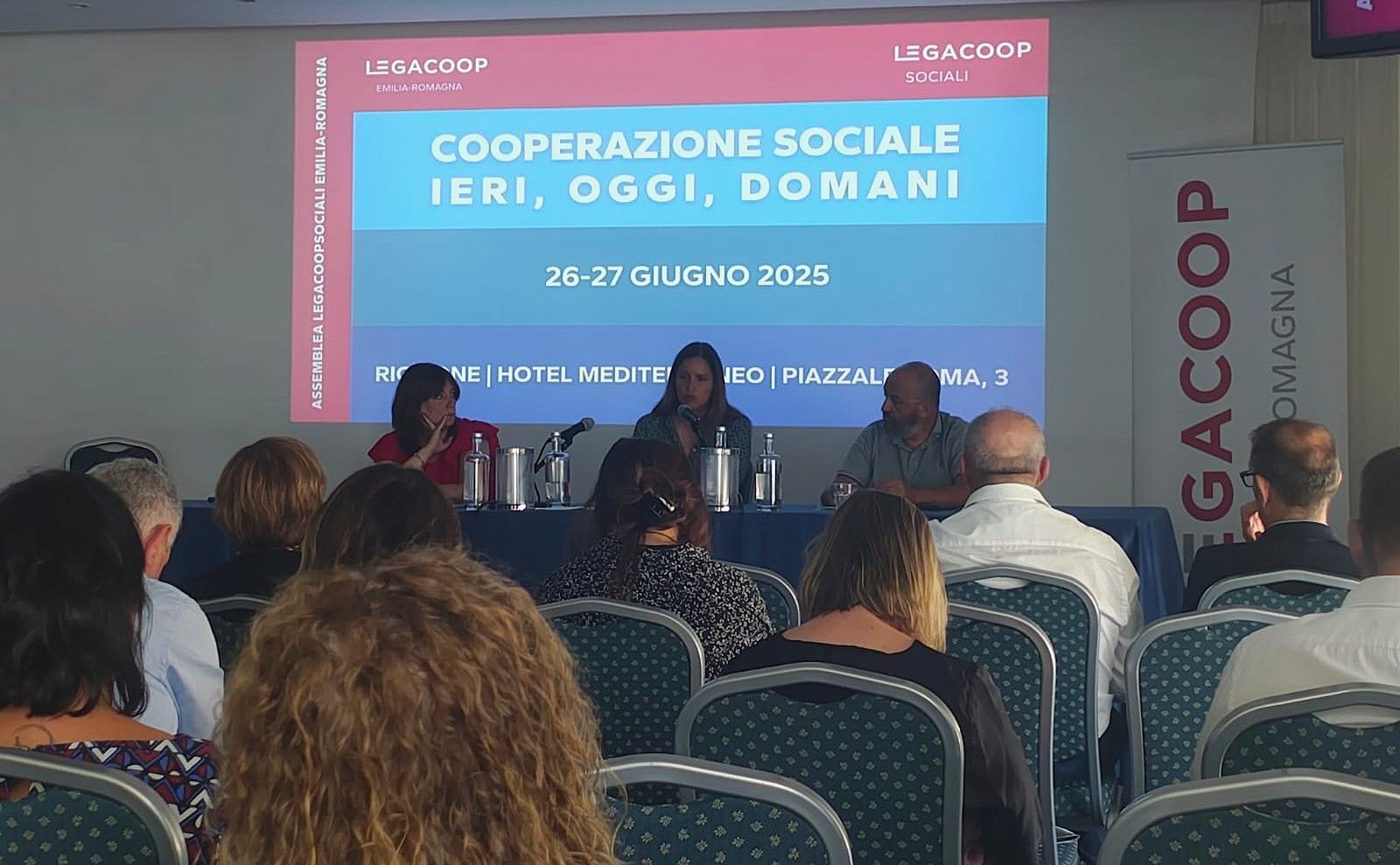 Tavola rotonda sulla cooperazione sociale durante una conferenza. Pubblico in primo piano, relatori seduti a un tavolo.