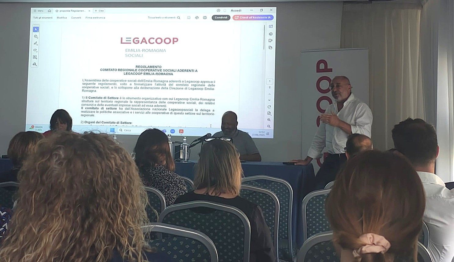 Un uomo tiene una presentazione a una conferenza. Il testo è sullo schermo del proiettore; i partecipanti sono seduti ai tavoli.
