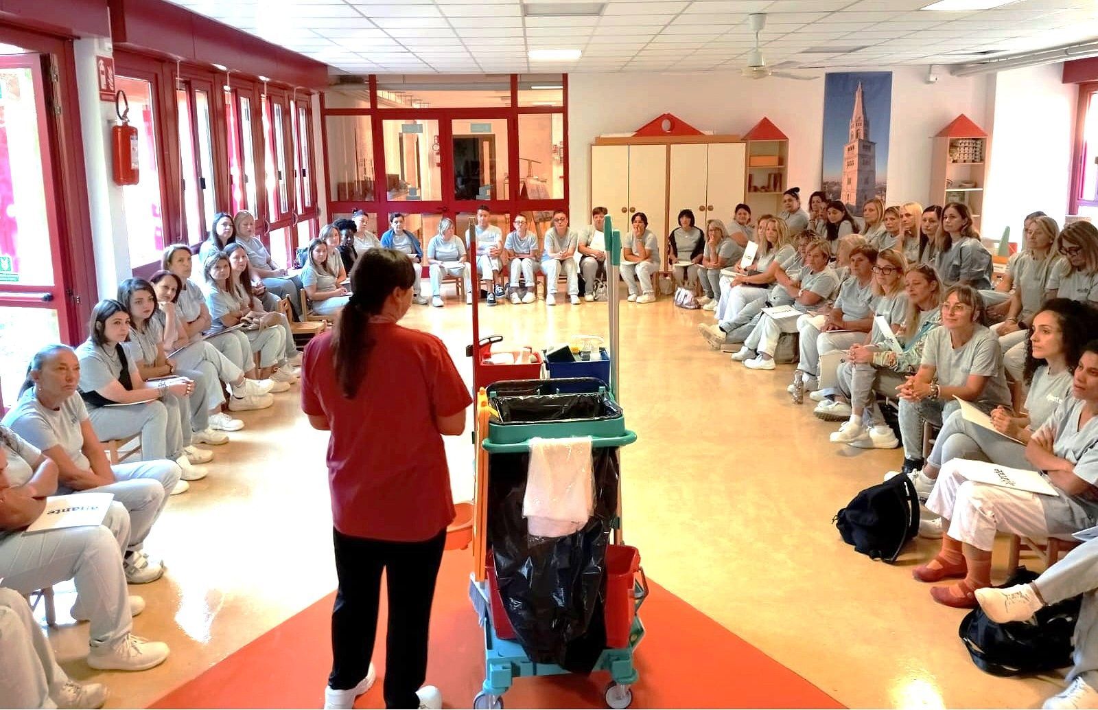 Una persona con una maglietta rossa mostra l'attrezzatura per la pulizia a un gruppo di persone sedute in una stanza ben illuminata.