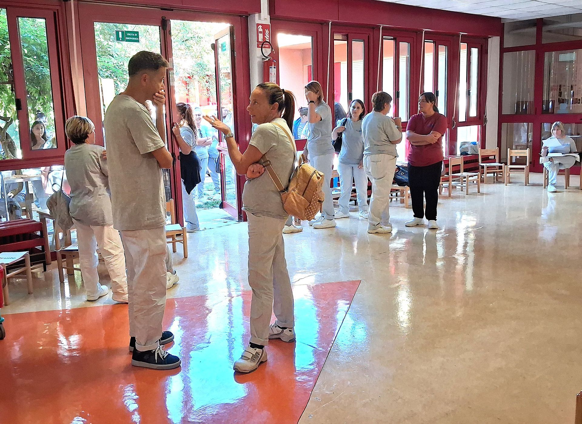 Due persone che parlano in un interno, altre sullo sfondo, vicino a porte aperte. Stanza ben illuminata con accenti rossi.