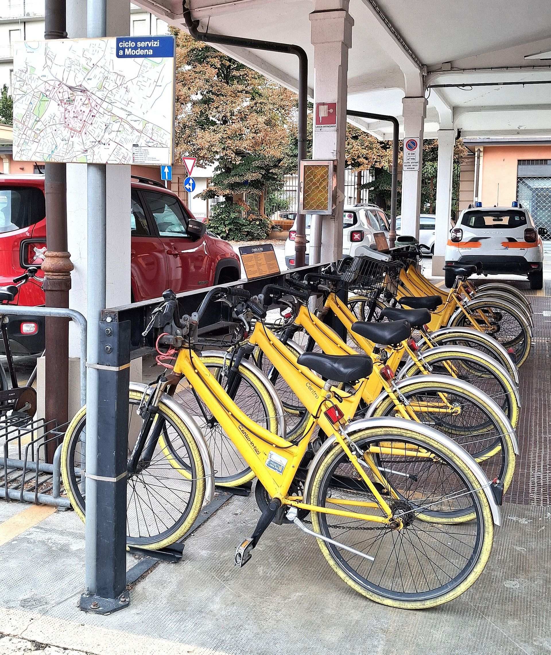Biciclette gialle parcheggiate in fila presso una stazione di bike sharing coperta. Nelle vicinanze sono visibili una mappa e alcune auto.