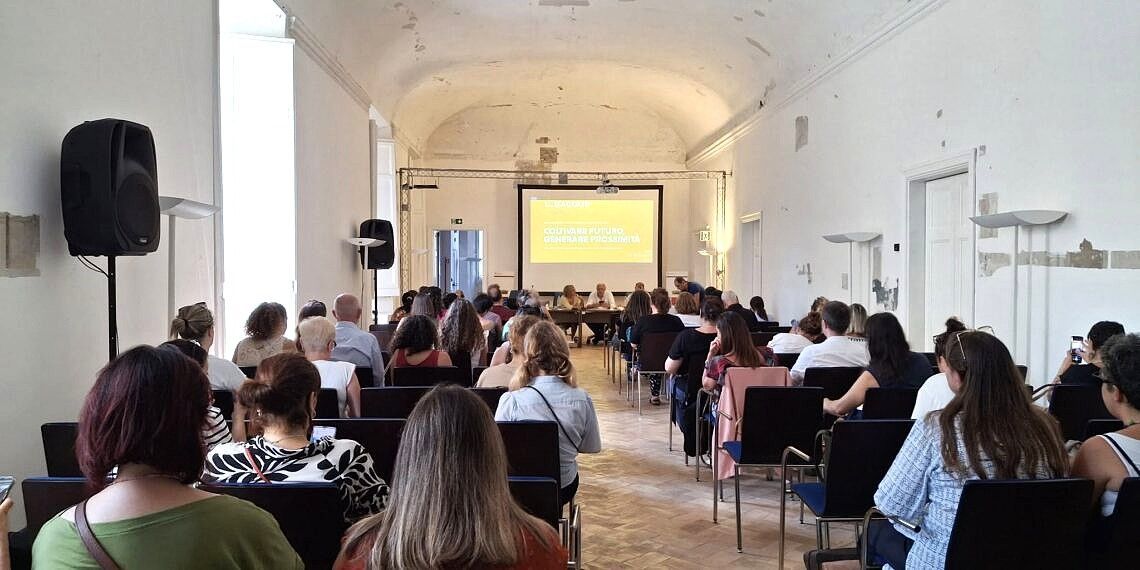 Il pubblico è seduto in una grande sala conferenze bianca, di fronte a uno schermo con testo. I relatori sono in prima fila.