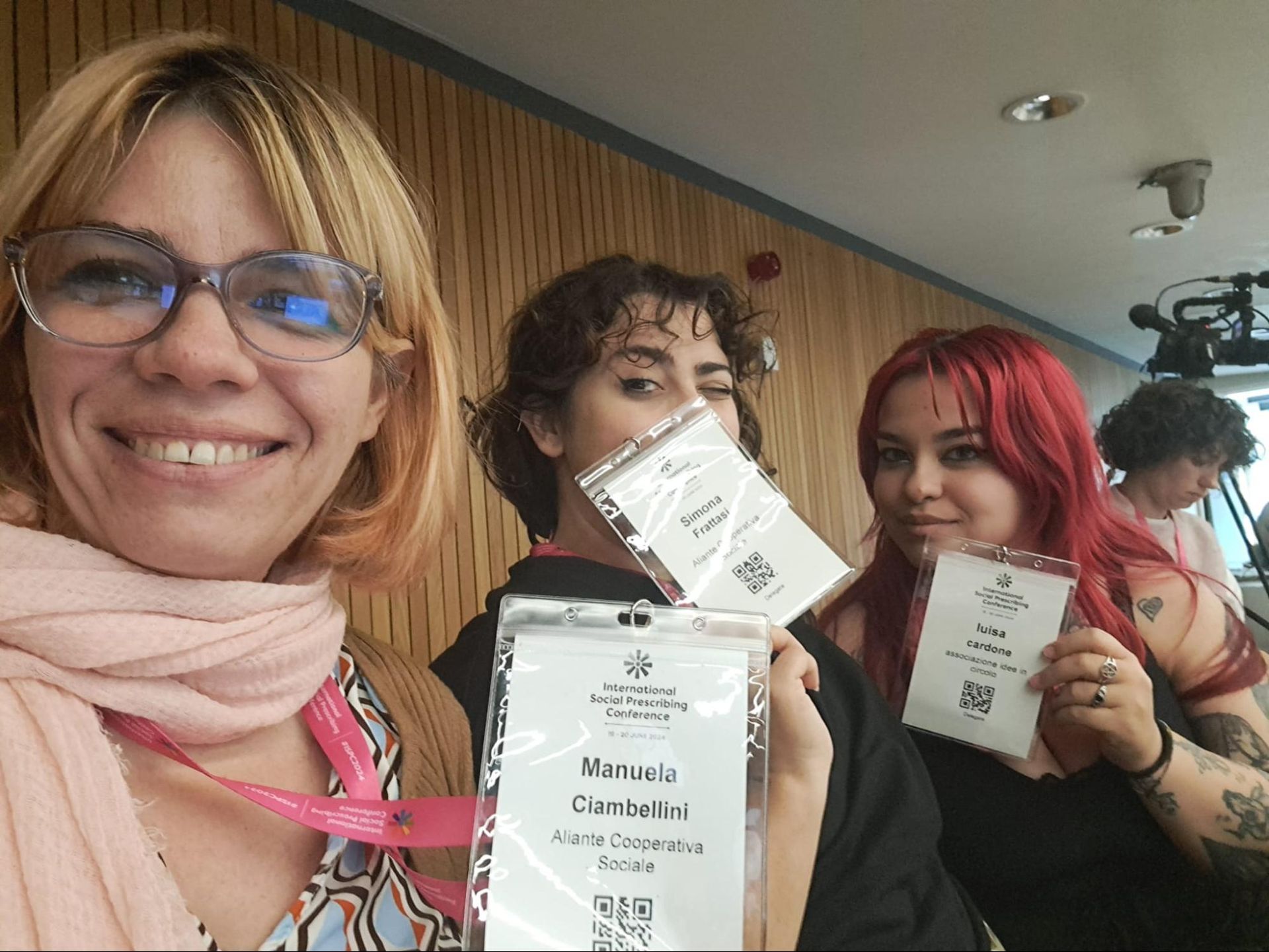 Tre persone sorridenti con cartellini identificativi. Una donna con gli occhiali si scatta un selfie. Ambiente per conferenza.