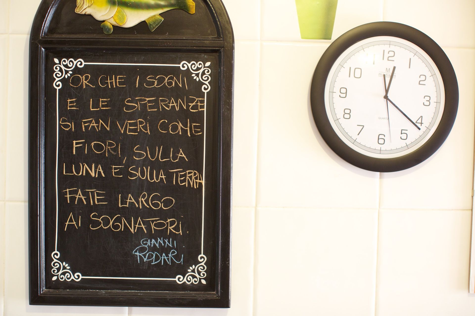 Una lavagna con scritte in italiano e un orologio su una parete piastrellata.