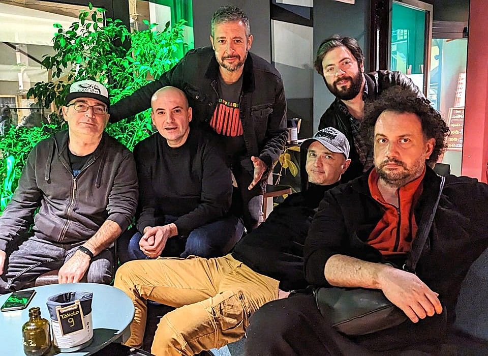 Un gruppo di sei uomini posa per una foto all'aperto. Uno si sporge in avanti con il braccio teso, gli altri sorridono.