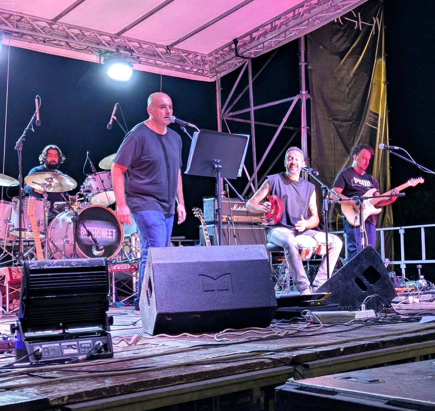 La band si esibisce sul palco di notte; cantante, chitarrista, batterista, percussionista; il palco è illuminato con luci rosa.