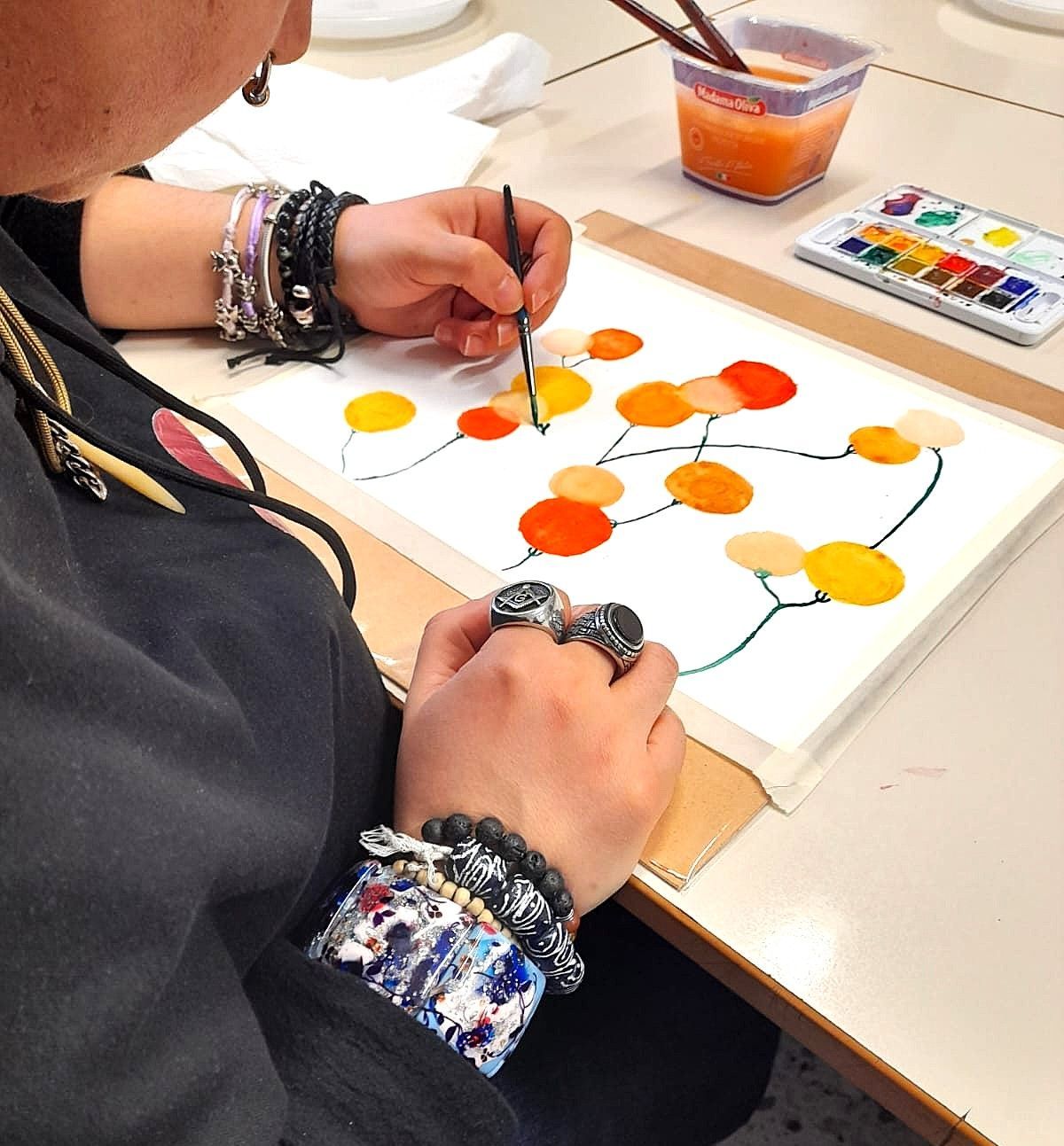 Persona che dipinge un disegno astratto colorato con gli acquerelli. Artista che indossa braccialetti e lavora a un tavolo con colori e una tazza d'acqua.