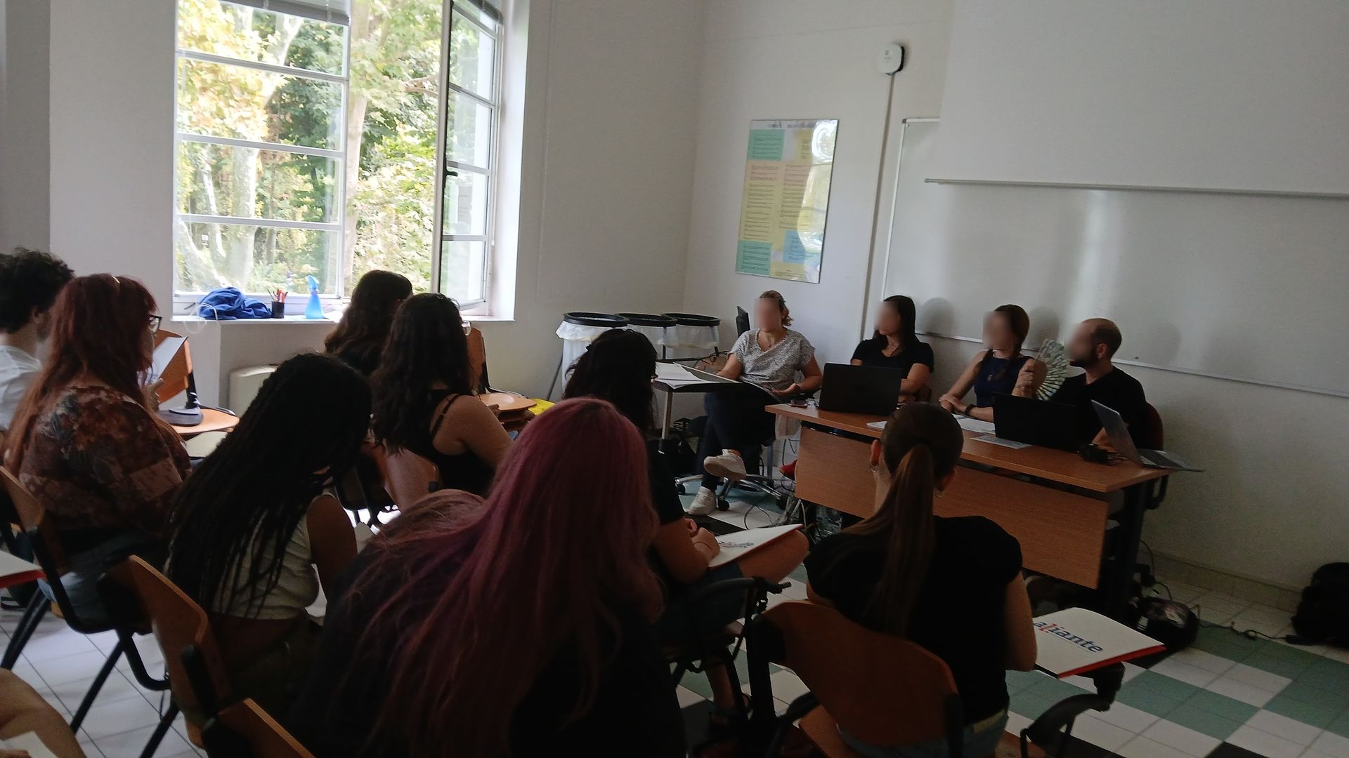 Un'aula scolastica, studenti che ascoltano una presentazione. Quattro persone sono sedute a un tavolo davanti a una lavagna.