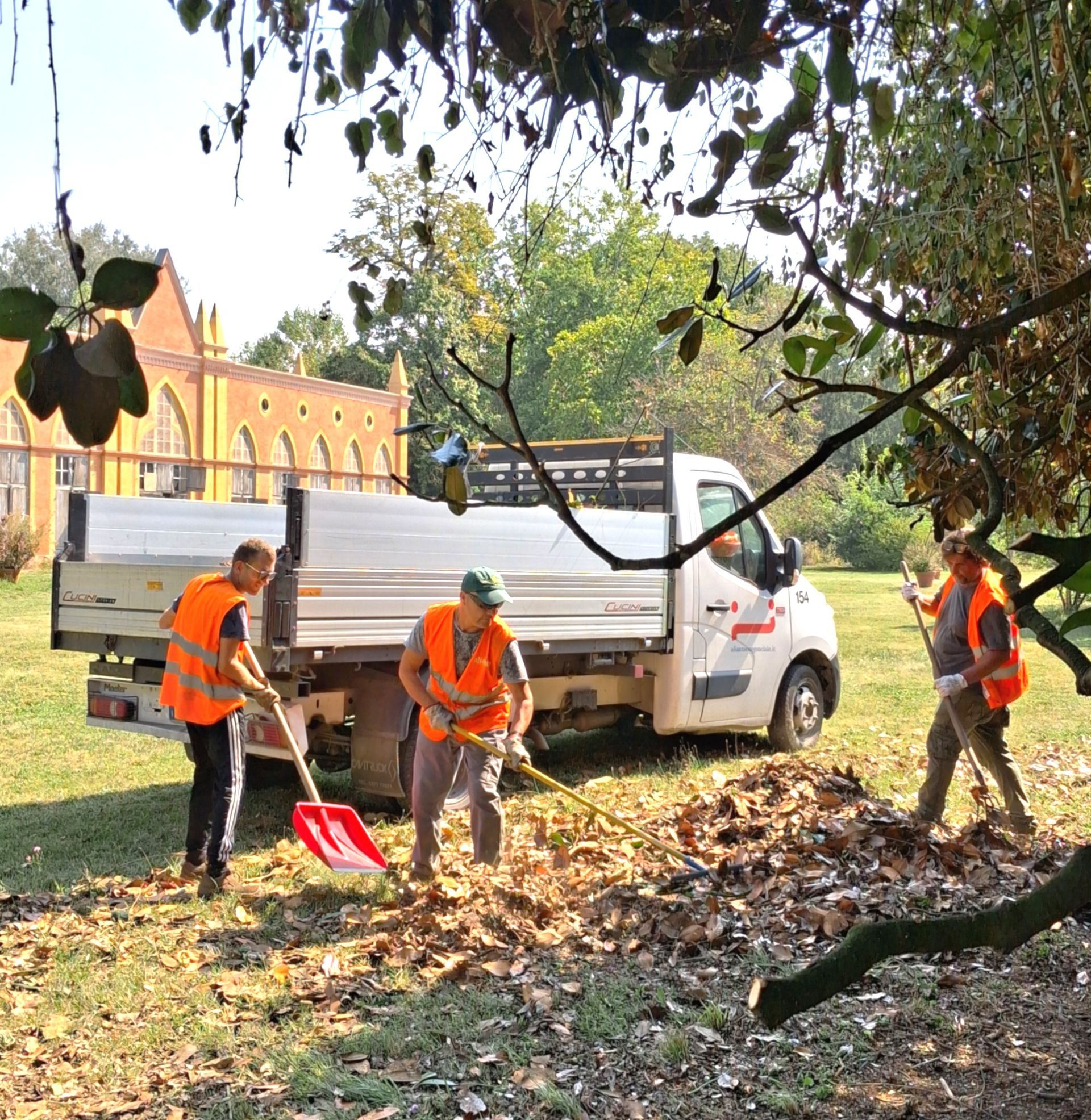 Tre lavoratori con i giubbotti arancioni rastrellano le foglie vicino a un camion in un parco, con un edificio sullo sfondo.