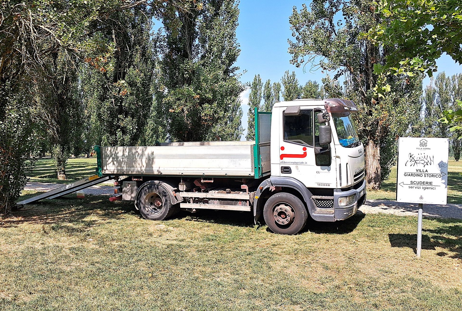 Un camion bianco con cassone ribaltabile parcheggiato sull'erba, accanto agli alberi. Una rampa metallica si estende dal cassone del camion.