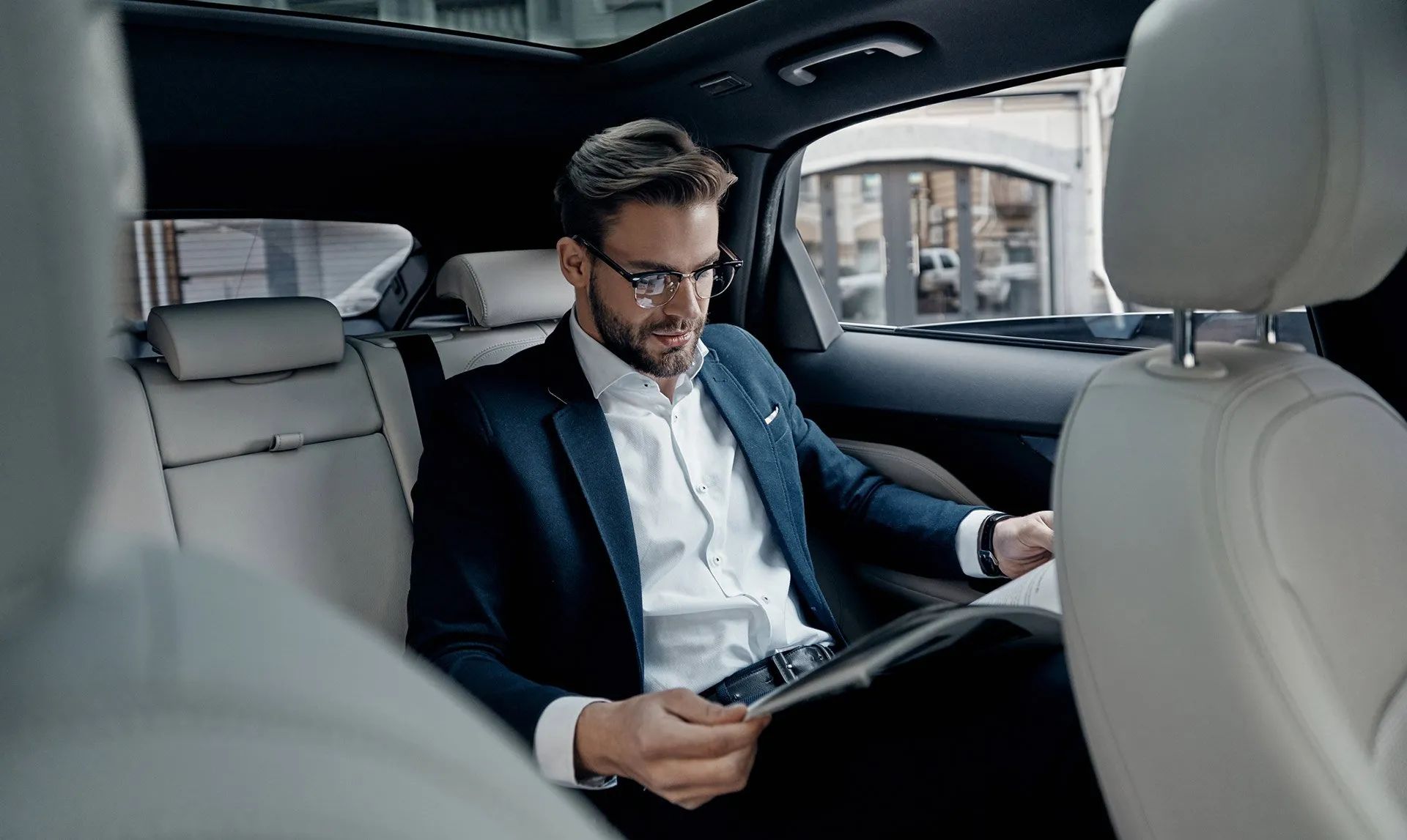 A man in a suit is sitting in the back seat of a car reading a magazine.