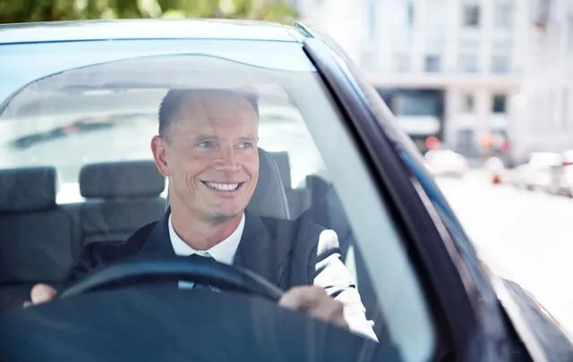 A man in a suit and tie is driving a car and smiling.