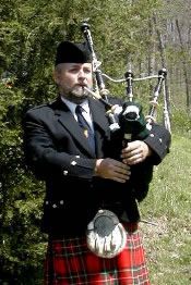 A person in a kilt and black jacket plays a set of bagpipes outdoors in front of a tree.