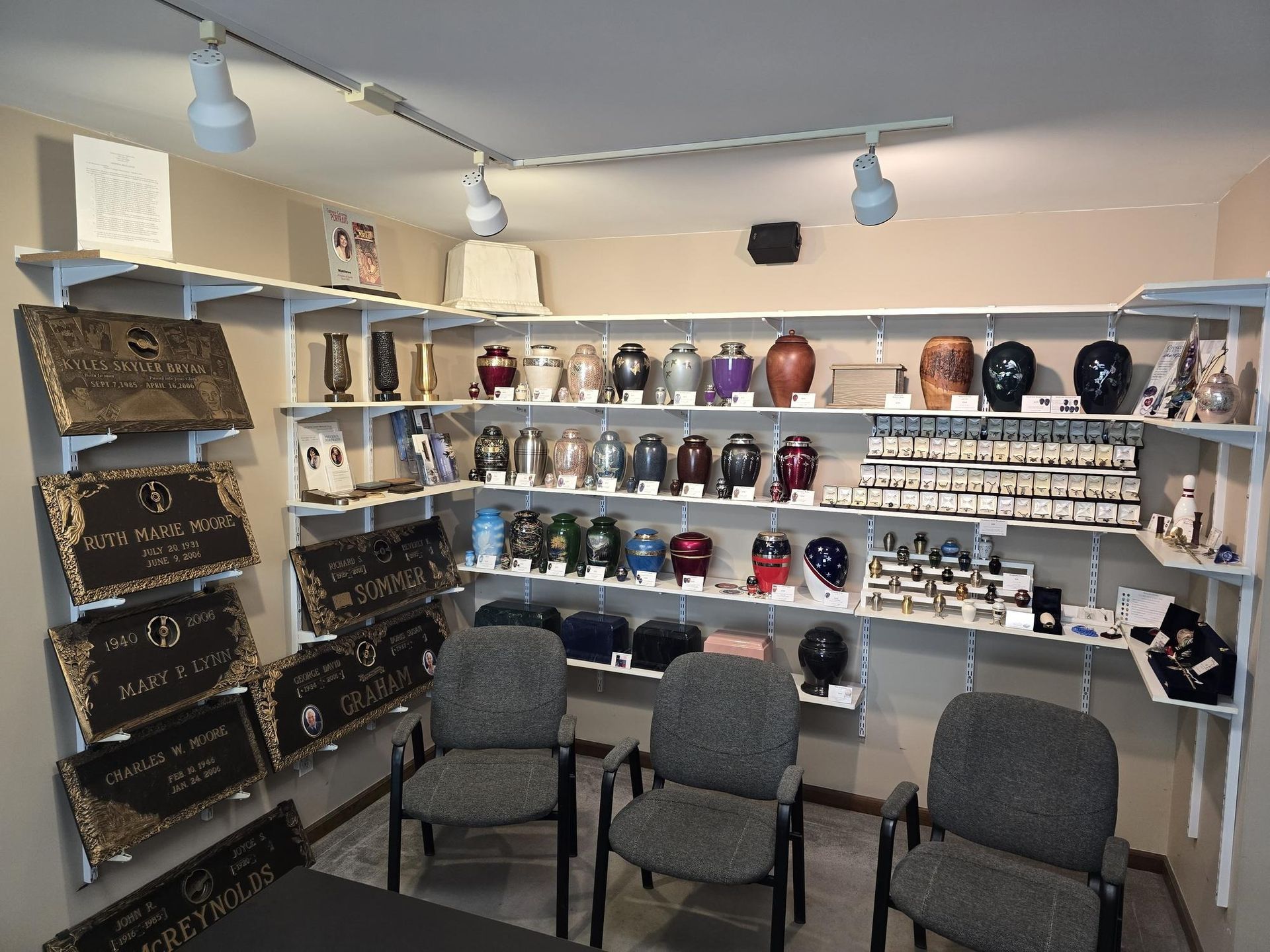 A display room with shelves containing rows of cremation urns and memorial plaques, with three grey chairs in front.