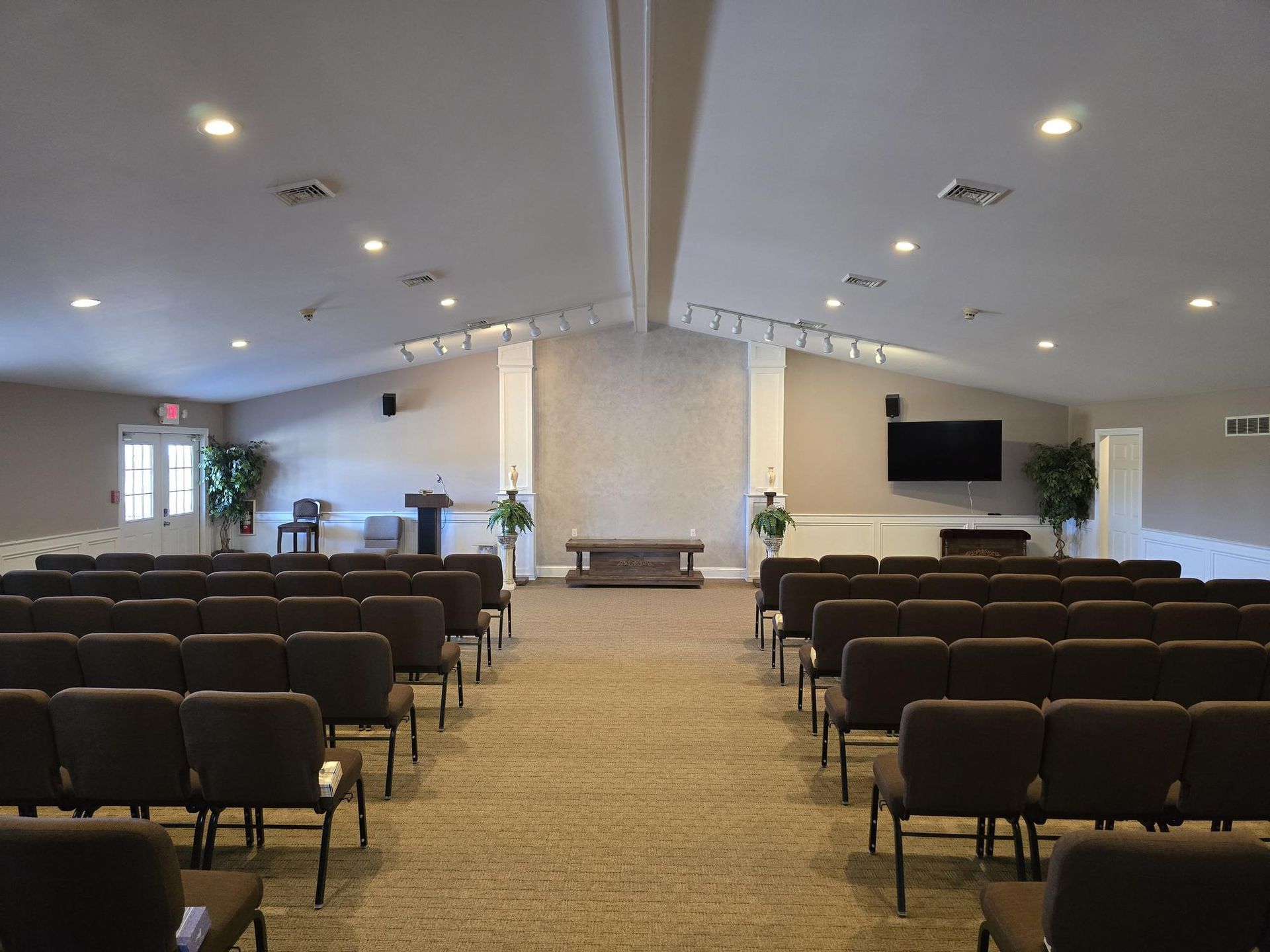 Interior of a church or community hall with rows of brown chairs facing a central altar and a wall-mounted TV.