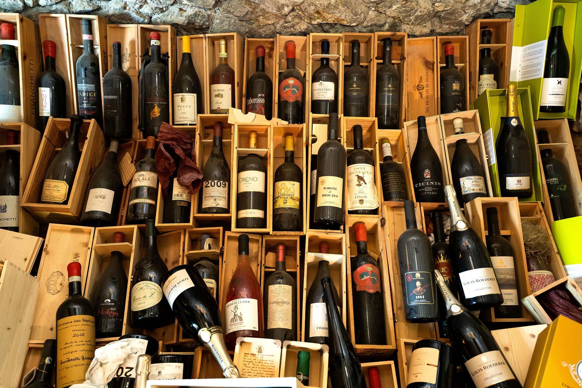 Diverse bottiglie di vino esposte in casse di legno su scaffali, creando una scena colorata che ricorda un mercato o una cantina.