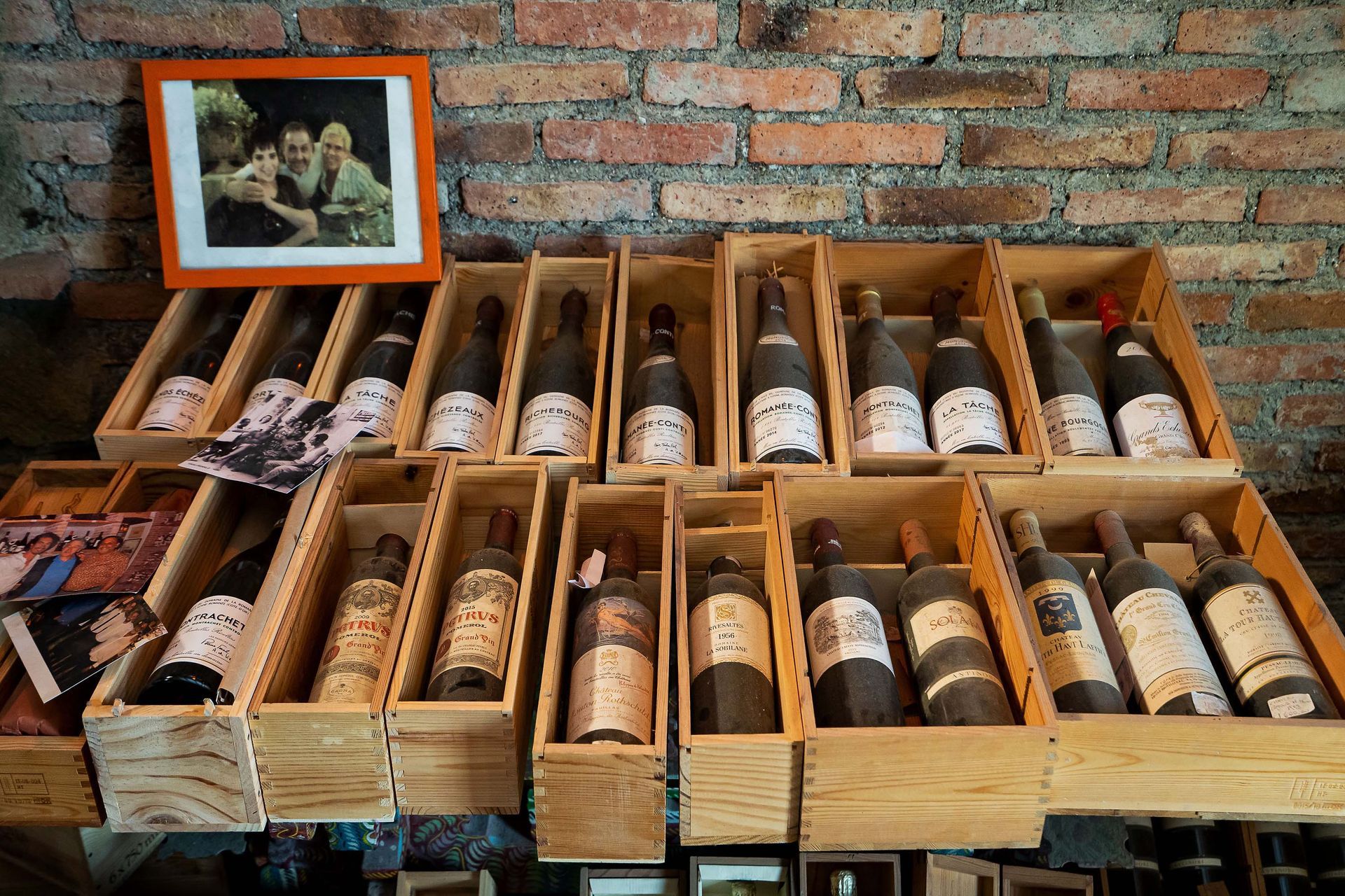 Espositore in casse di legno con diverse bottiglie di vino accanto a una foto incorniciata su un muro di mattoni.