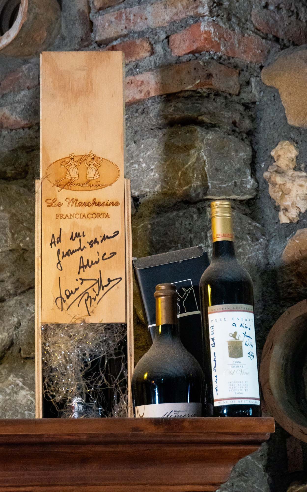 Bottiglie di vino e una bottiglia alta in scatola esposte su uno scaffale di legno appoggiato a un muro di pietra.