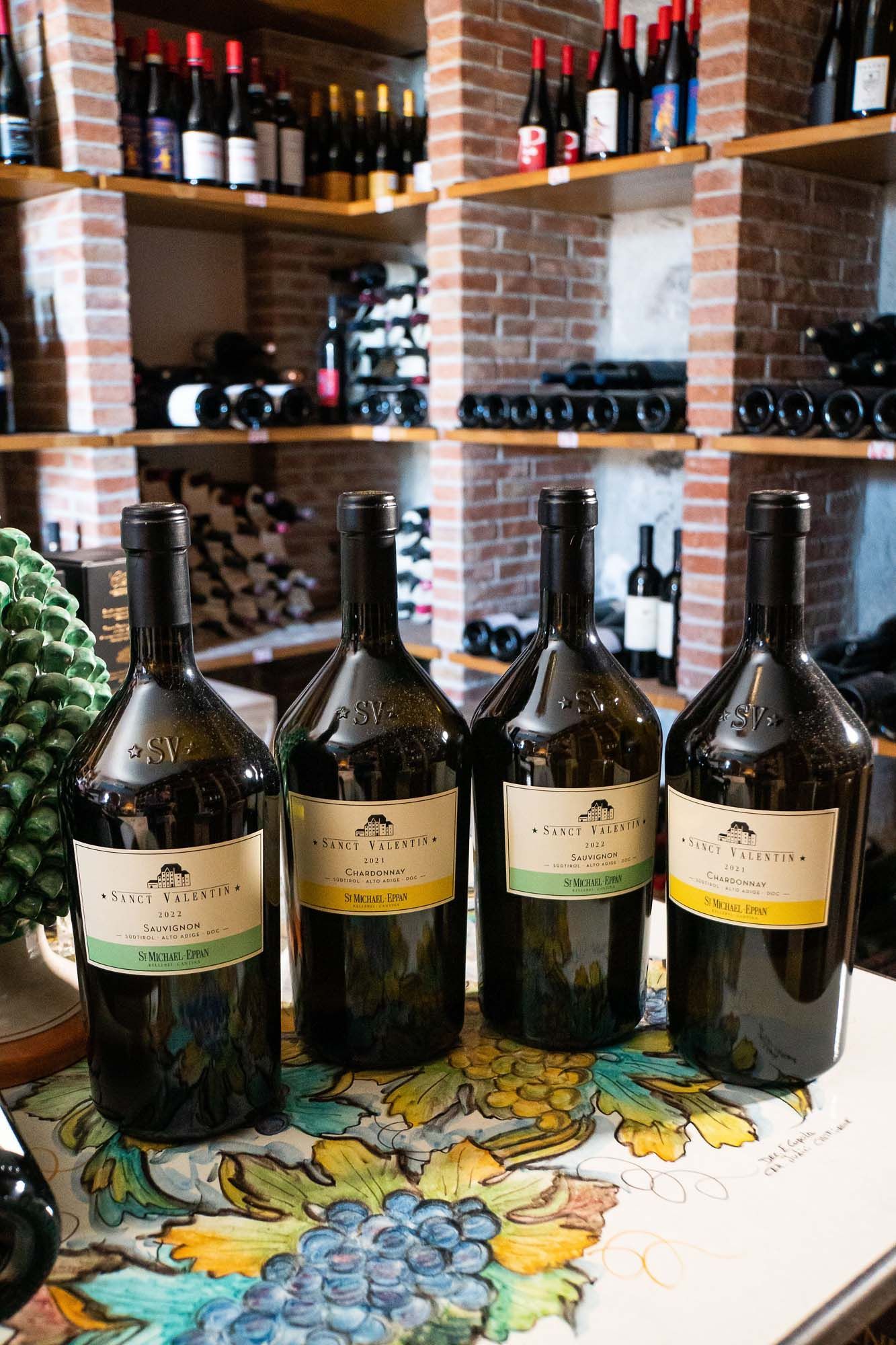 Quattro grandi bottiglie di vino su un tavolo in una cantina con pareti in mattoni, dietro la quale si trovano scaffali pieni di bottiglie.