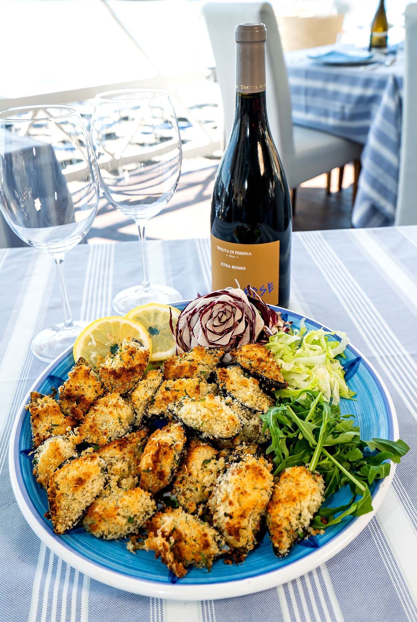 Un piatto blu di frutti di mare fritti impanati con limone e insalata su un tavolo in riva al mare, con una bottiglia di vino sullo sfondo.