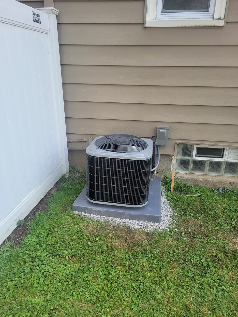 Outdoor air conditioner unit on a concrete pad beside tan house siding and a white fence.