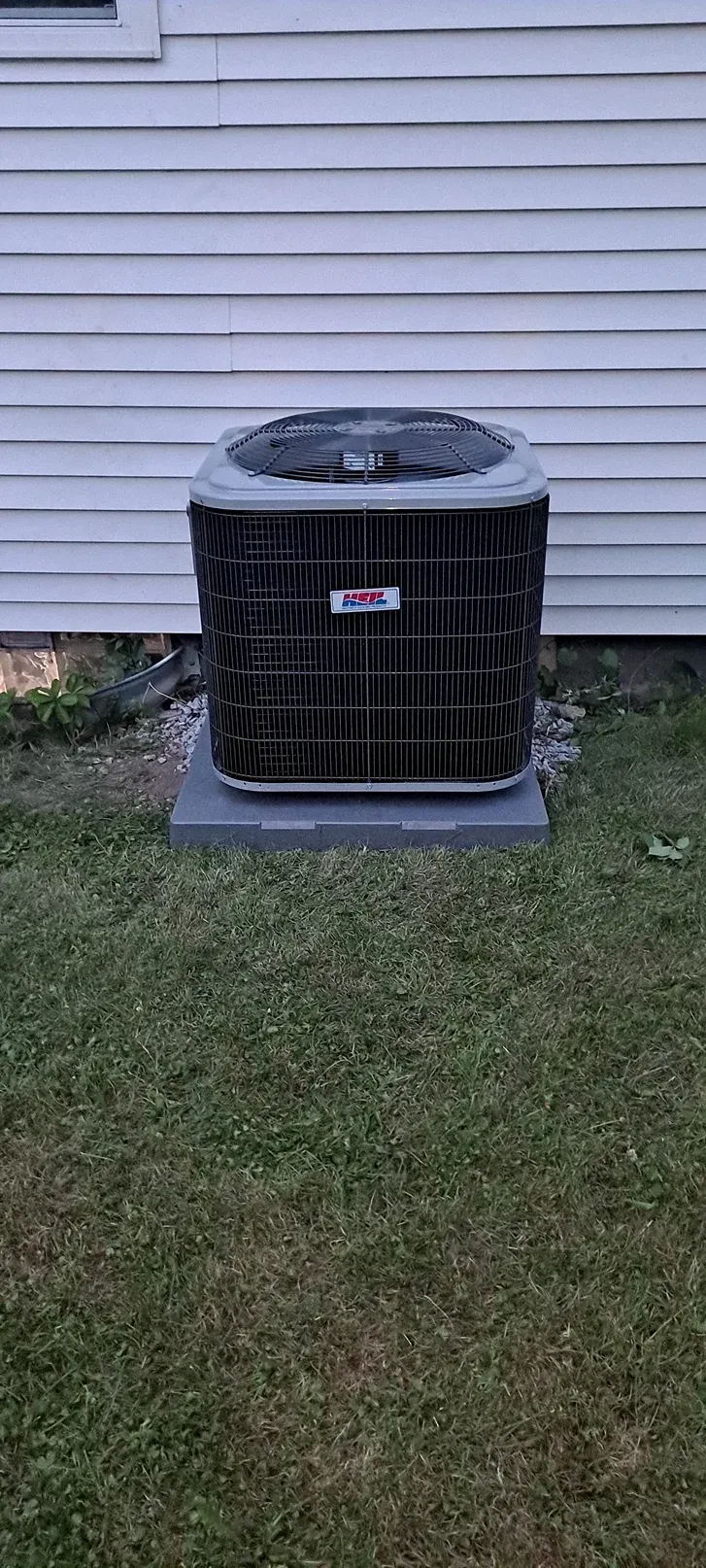 Outdoor air conditioner unit beside a house with white siding and a small grass lawn.