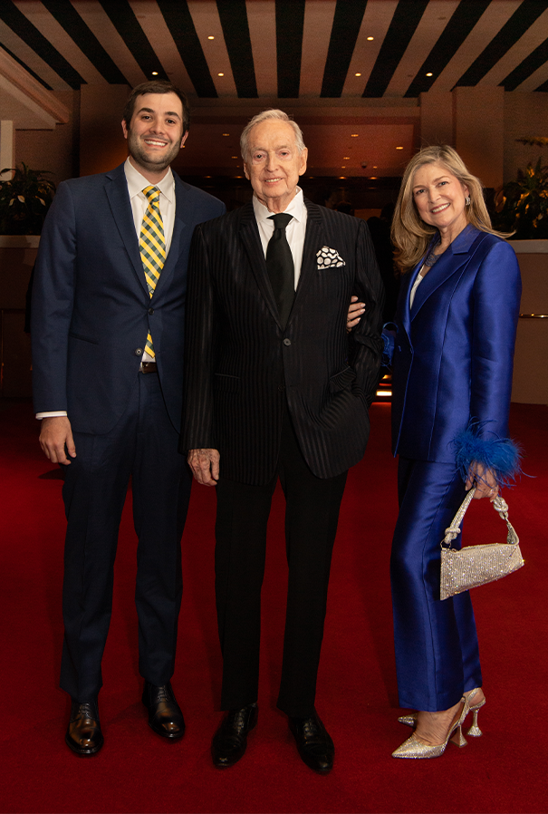 Three people pose on a red carpet: a man in a black suit, and a woman in blue, both with arms around the man in the middle.