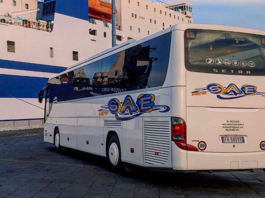 Pullman turistico bianco con grafiche blu e arancioni parcheggiato accanto a una grande nave in banchina