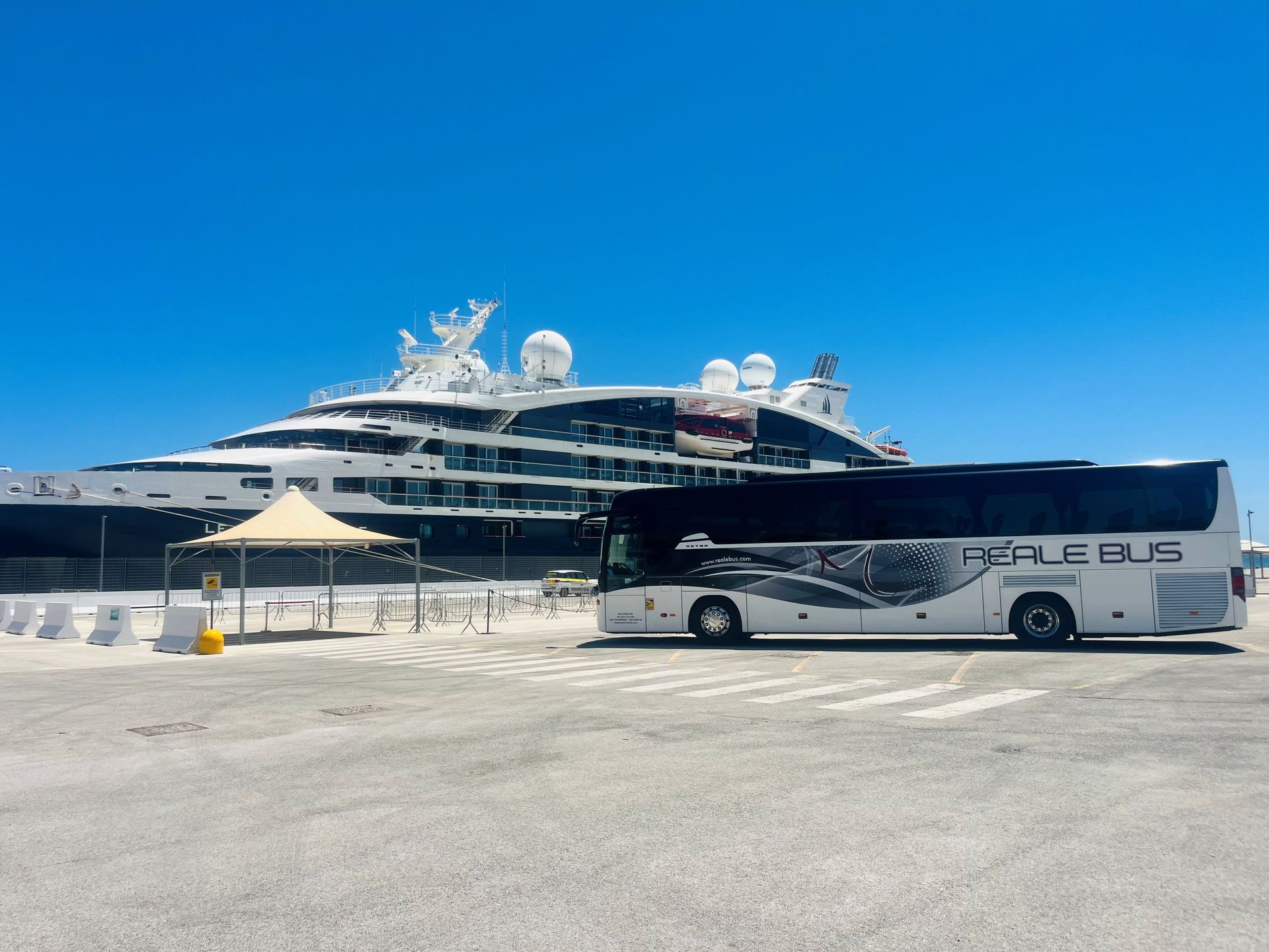 Autobus turistico bianco parcheggiato accanto a una grande nave da crociera in un porto soleggiato, sotto un cielo azzurro e limpido