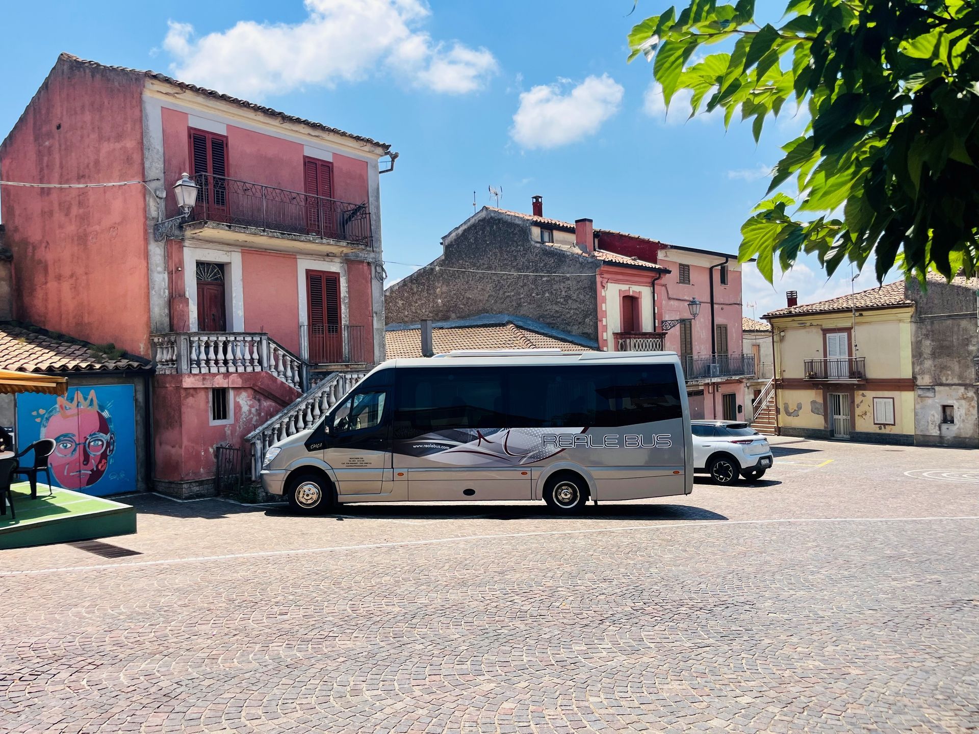 Un minibus argentato parcheggiato in una soleggiata piazza acciottolata, circondata da edifici dai colori pastello e sovrastata da un cielo azzurro.
