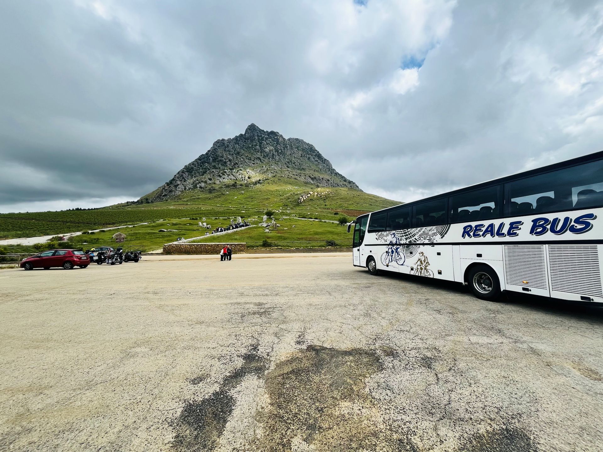 Autobus parcheggiato accanto a un piazzale di ghiaia ai piedi di una montagna, sotto un cielo nuvoloso