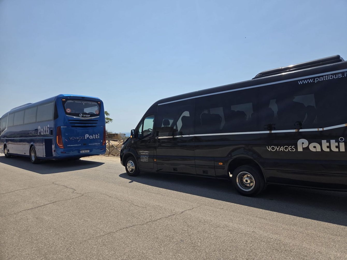 Due pullman parcheggiati, un autobus blu e un furgone nero, in un piazzale soleggiato e aperto
