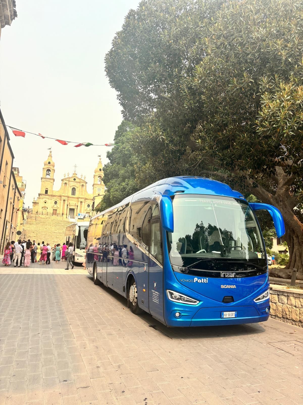 Autobus turistico blu su una strada acciottolata, accanto a una chiesa storica e a grandi alberi