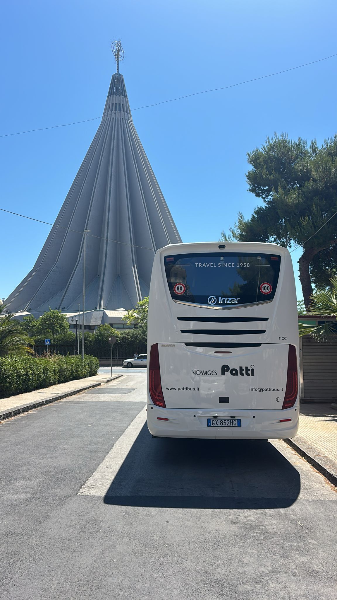 Autobus bianco su una strada, di fronte a un alto monumento moderno bianco, sotto un cielo azzurro e terso.