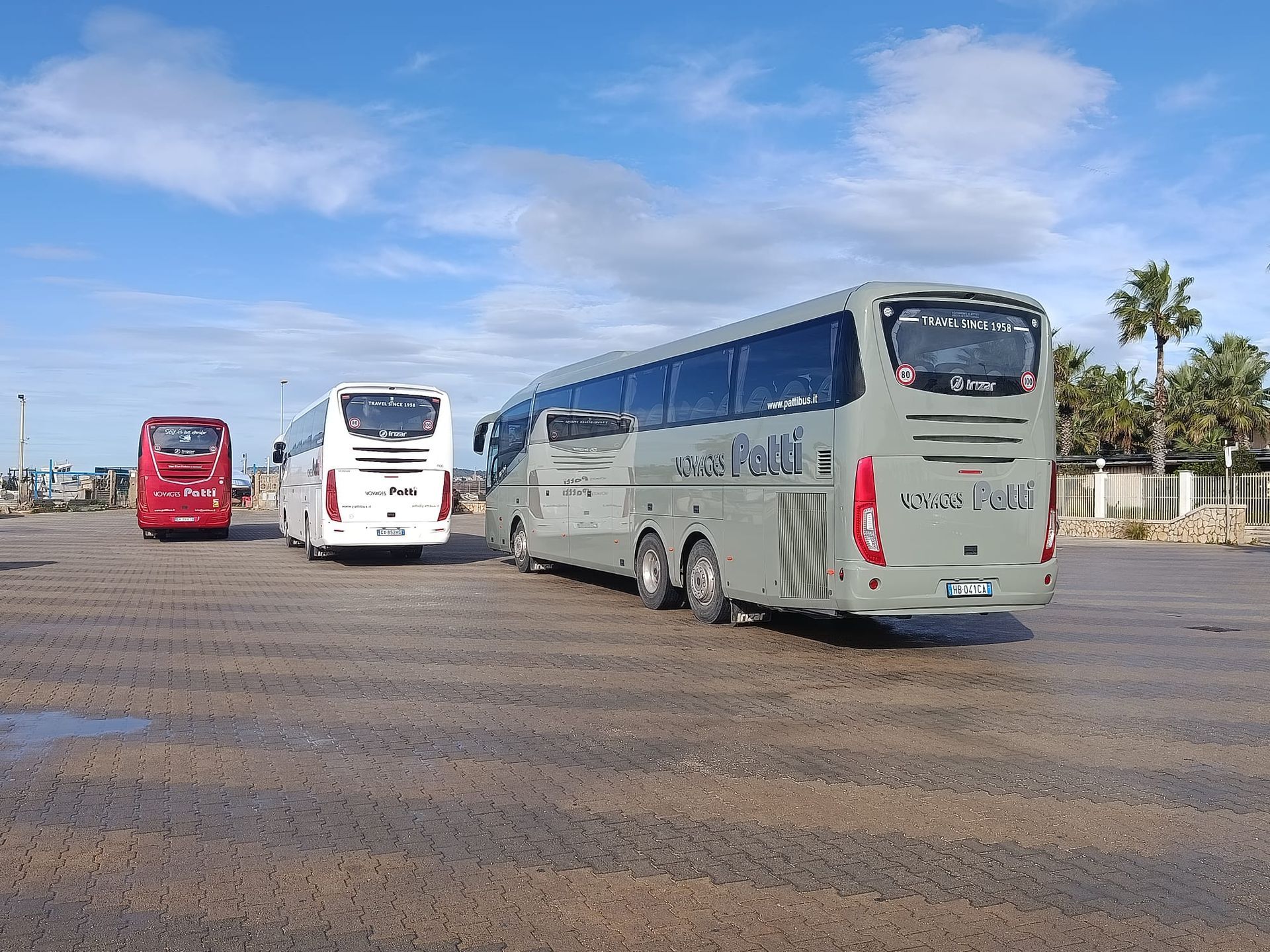 Tre autobus parcheggiati in un ampio piazzale sotto un cielo azzurro, tra cui pullman rossi, bianchi e grigi.