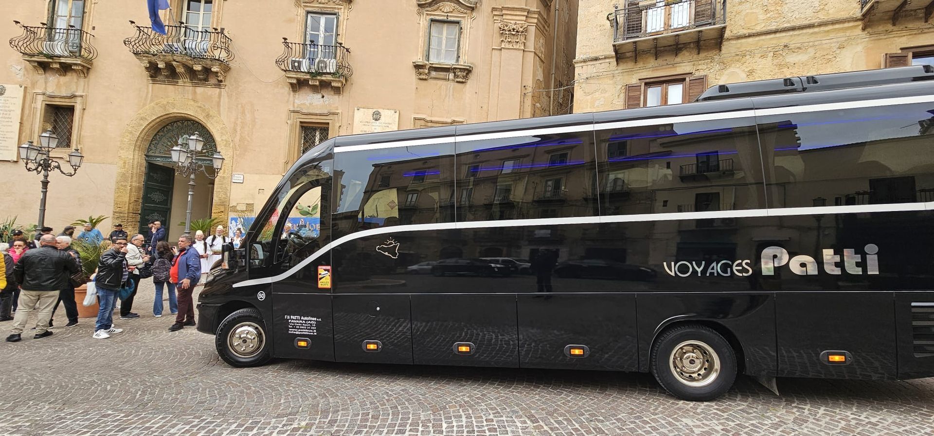 Autobus nero parcheggiato davanti a un edificio in pietra, con alcune persone riunite nelle vicinanze.