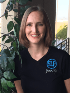 Smiling person in a black SLP T-shirt standing beside a leafy plant indoors